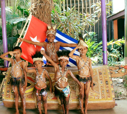 West Papuan boys in traditional dress, saluting in front of their national flag and a garamut drum 